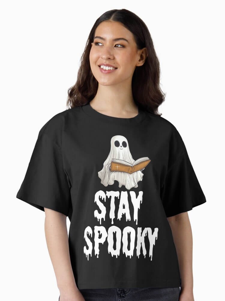 Person wearing a black boxy ‘Stay Spooky’ t shirt with a cute ghost reading a book and dripping white lettering.
