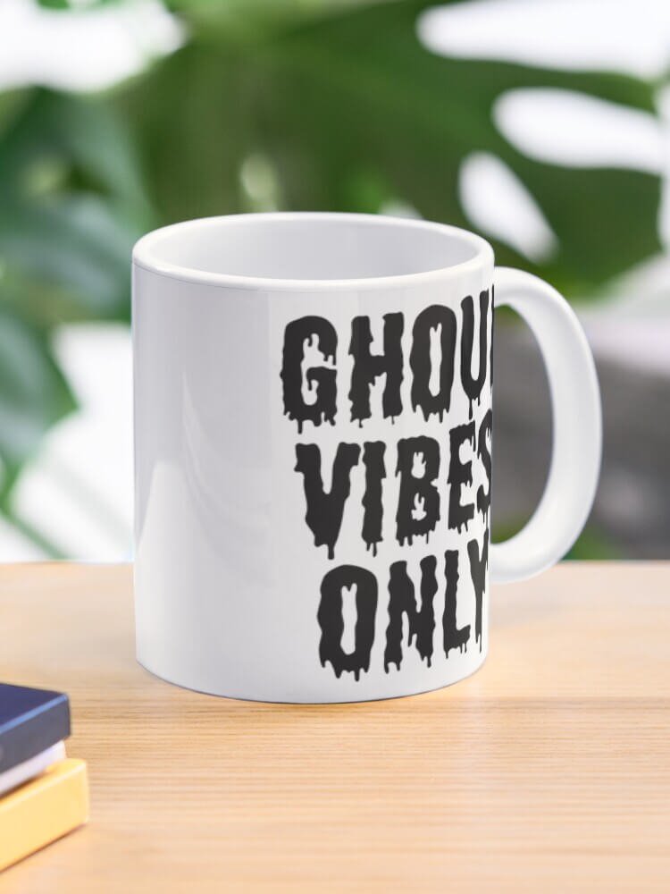 White ceramic mug on a wooden desk with bold black dripping text reading ‘Ghoul Vibes Only.