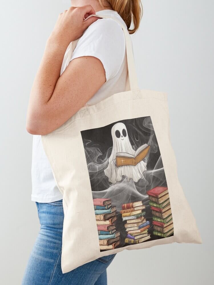 Person carrying a natural cotton bookish spirit tote bag with a ghost reading and book stacks on the front
